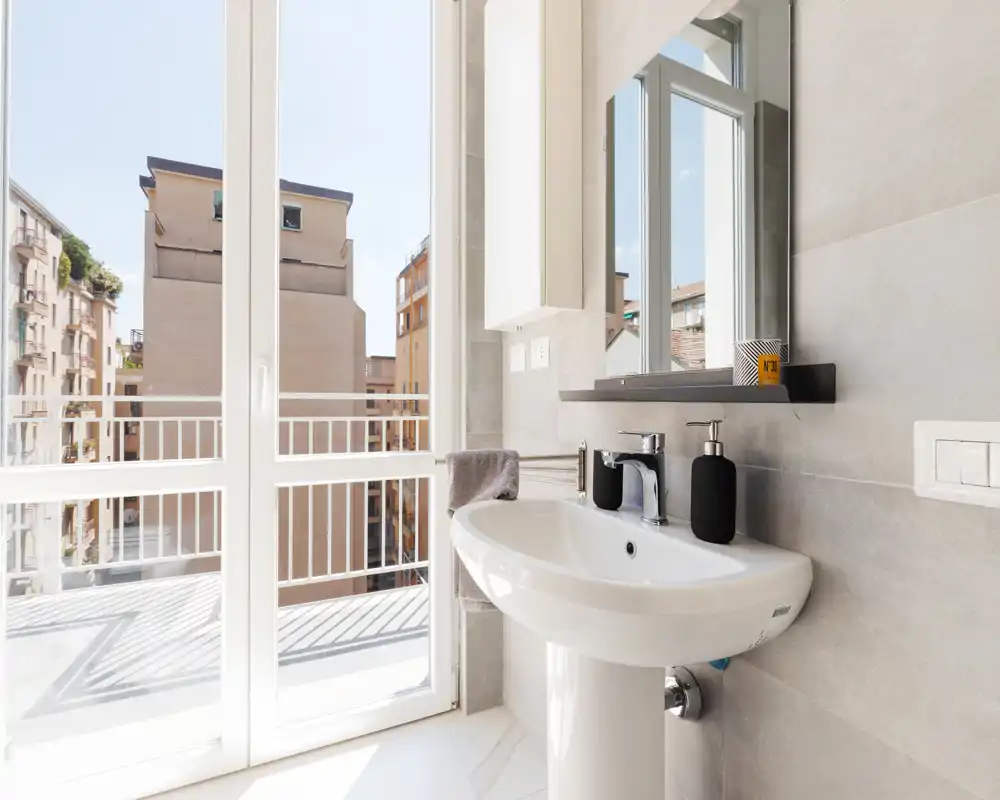 Bright modern bathroom with a pedestal sink, wall-mounted mirror and large balcony/terrace doors that flood the space with natural light.