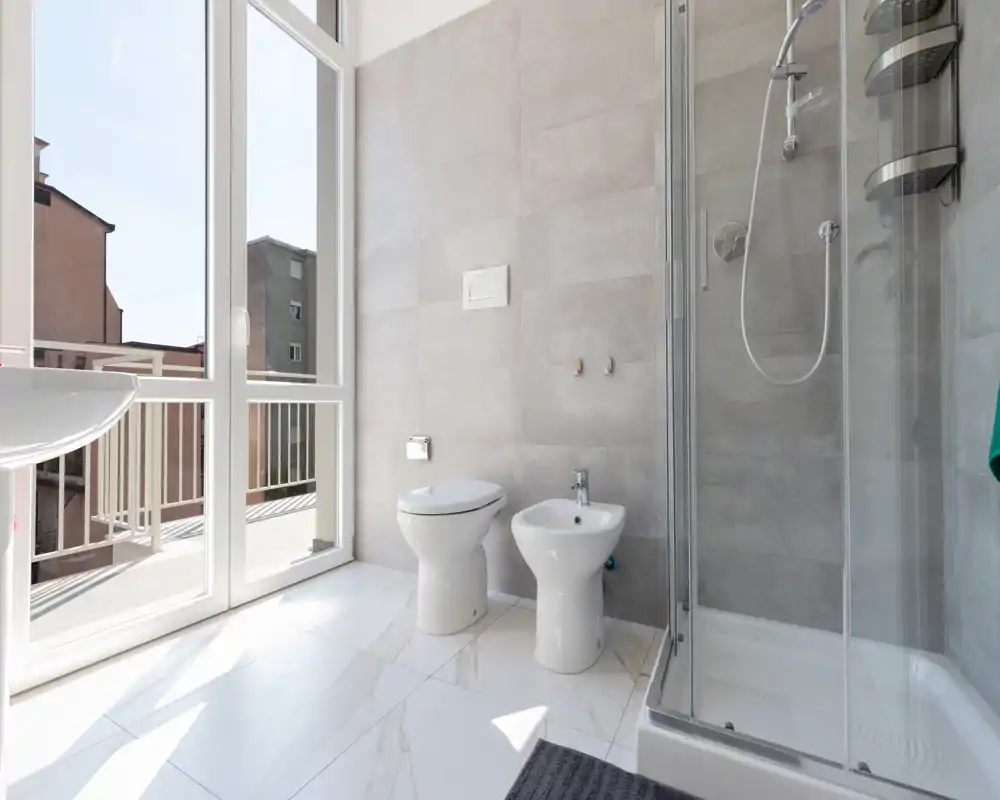 Bagno moderno e luminoso con box doccia, WC e bidet, ampie finestre che si affacciano sul balcone; pavimento effetto marmo e rivestimenti neutri.