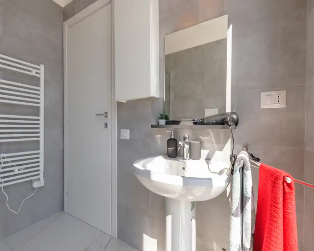 Bagno moderno e luminoso con lavabo a colonna, specchio a parete e scaldasalviette; piastrelle grigie neutre e luce naturale per un ambiente pulito e fresco.