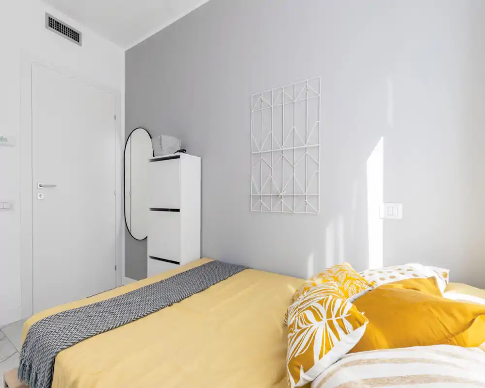 Camera da letto moderna e luminosa con un letto matrimoniale rifinito con biancheria gialla, cuscini decorativi e una coperta a motivi. Arredi minimali: mobiletto bianco alto, uno specchio ovale e una parete grigia chiara che riceve molta luce naturale.