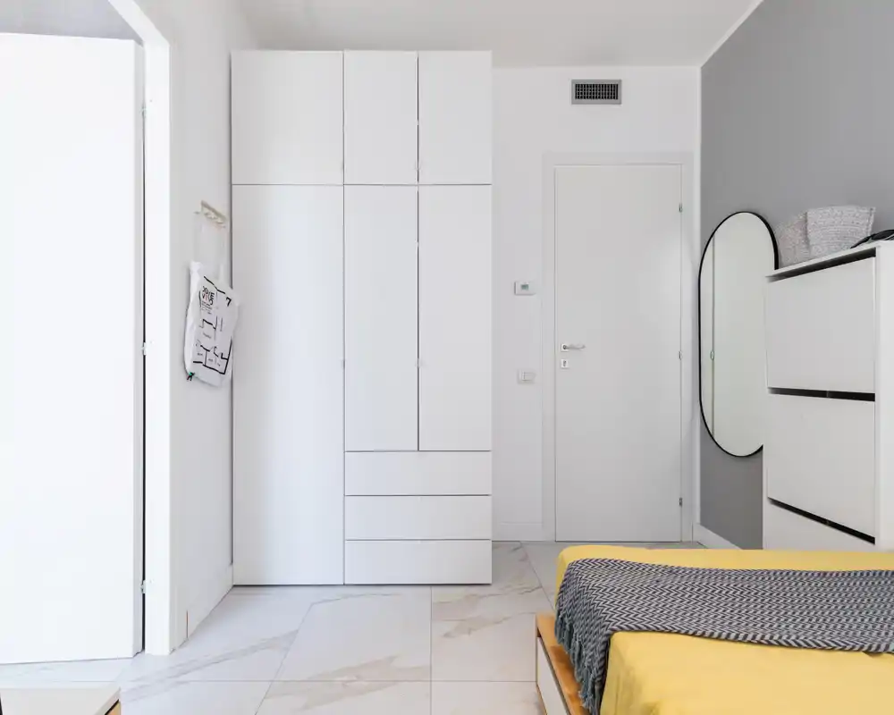 Angolo di camera da letto moderno e luminoso con letto basso in piattaforma con copriletto giallo e plaid, armadio bianco pulito, comò e specchio ovale a parete per un look minimalista.