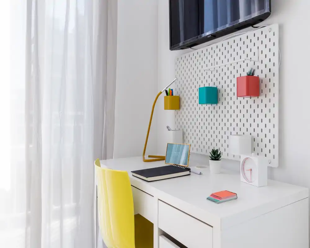 Area di lavoro luminosa e minimal vicino alla finestra con scrivania bianca, sedia gialla, lampada da tavolo e portaoggetti a parete — perfetta per studiare o lavorare da remoto.