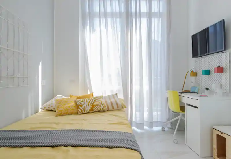 Camera singola moderna e luminosa con copriletto giallo, cuscini decorativi e una scrivania compatta sotto la TV a parete—luminoso e ben arredato, perfetto per un annuncio.