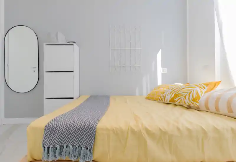 Camera da letto luminosa e moderna con un ampio letto con biancheria gialla, cuscini decorativi e una coperta intrecciata; pareti neutre, uno specchio alto e un mobiletto sottile completano lo stile minimal.