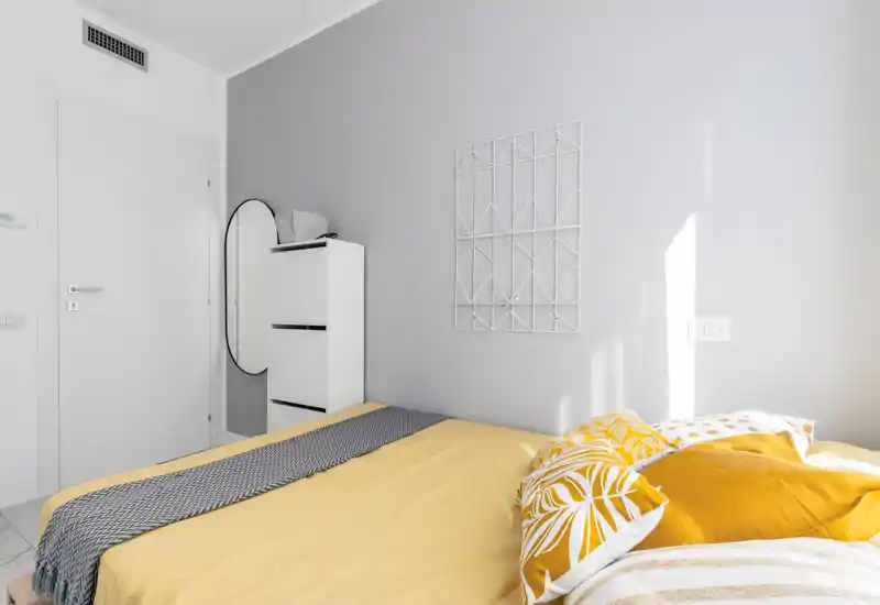 Camera da letto moderna e luminosa con un letto matrimoniale rifinito con biancheria gialla, cuscini decorativi e una coperta a motivi. Arredi minimali: mobiletto bianco alto, uno specchio ovale e una parete grigia chiara che riceve molta luce naturale.