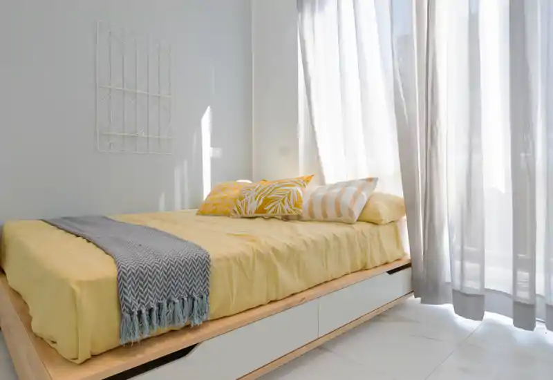 Camera da letto luminosa e ben allestita con letto basso in legno, biancheria gialla e cuscini decorativi; ampie tende leggere filtrano molta luce naturale.
