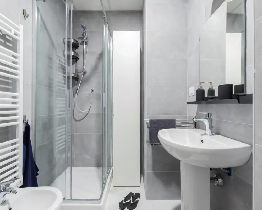Salle de bains moderne et compacte avec cabine de douche vitrée, lavabo sur colonne et carrelage gris neutre pour un aspect épuré et minimaliste.