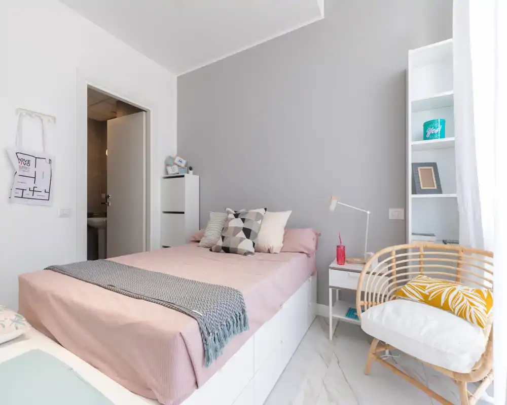 Chambre simple lumineuse et moderne avec lit bien fait, coussins décoratifs et fauteuil en rotin près de la fenêtre — idéale pour un séjour confortable.
