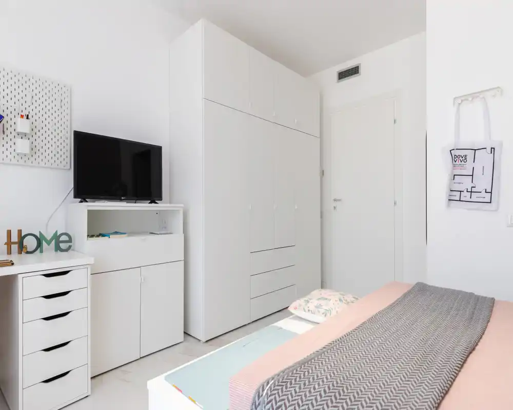 Chambre lumineuse et minimaliste avec lit en premier plan, grande armoire blanche et rangements soignés. Textiles doux et télévision complètent un agencement moderne, adapté à une annonce locative.