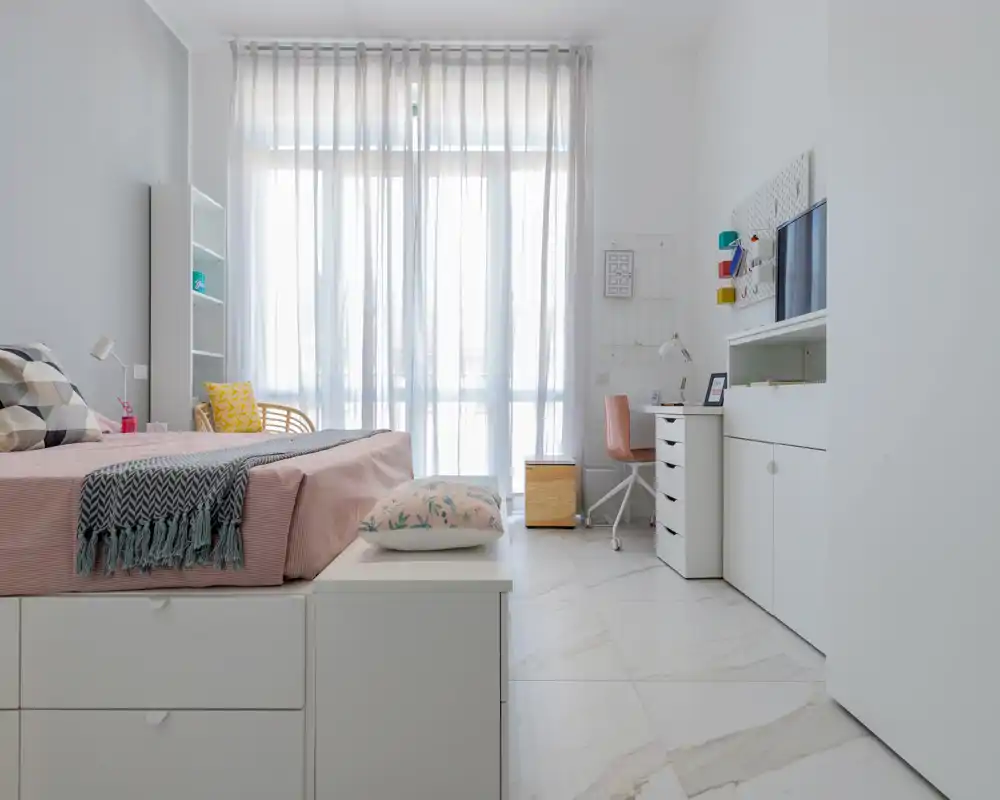 Chambre moderne et lumineuse avec lit bien fait, textiles doux et coffre de rangement en bout de lit ; grande fenêtre voilée apporte une belle lumière naturelle et un petit bureau est présent.