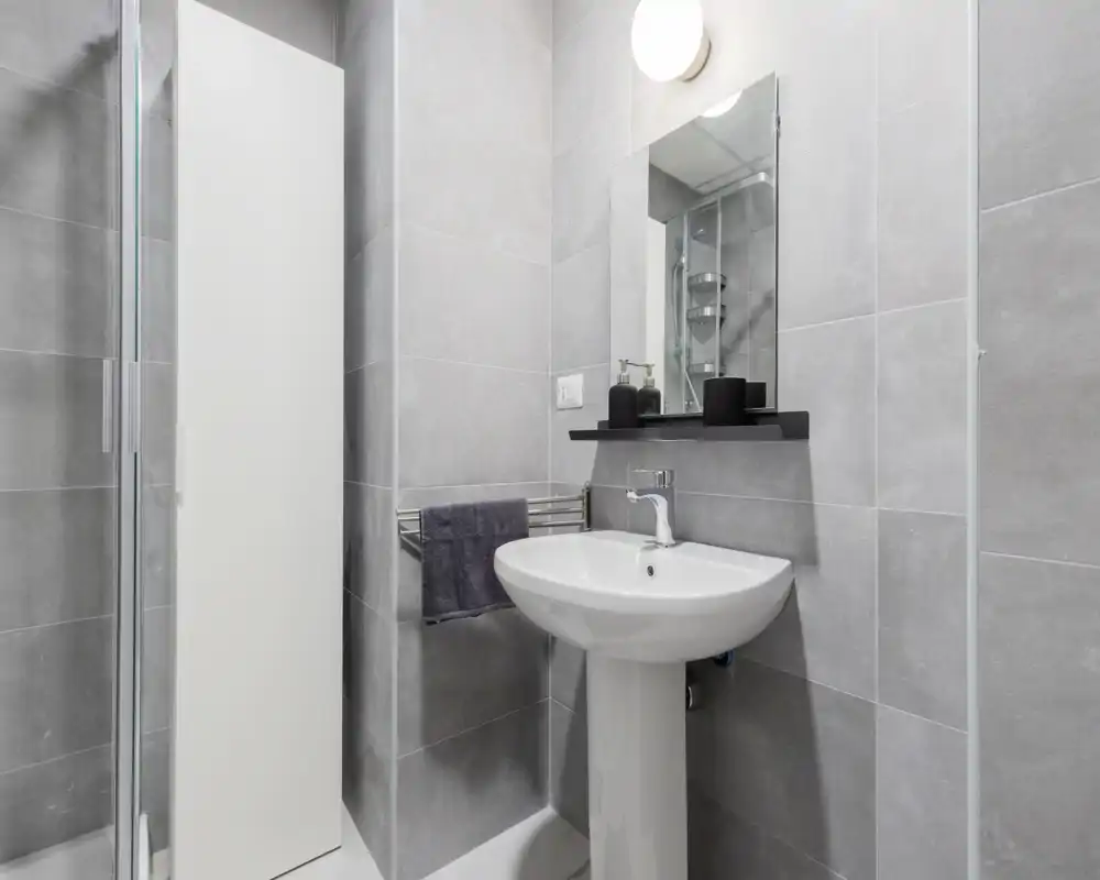 Salle de bains moderne et propre avec lavabo sur colonne, miroir, porte-serviettes et cabine de douche visible à gauche. Carrelage gris neutre et éclairage lumineux pour un rendu net et contemporain.