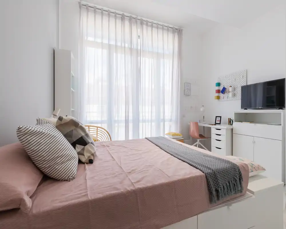 Chambre moderne et lumineuse avec un grand lit en linge rose, coussins décoratifs et plaid confortable. La pièce comprend un espace de travail avec bureau et meuble TV, et des voilages laissant entrer une belle lumière naturelle.
