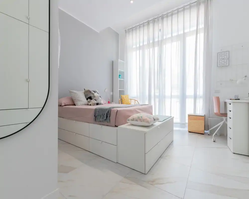 Chambre lumineuse et moderne avec lit sur base de rangement blanche, textiles pastel soignés et grandes voilages laissant entrer une belle lumière naturelle.