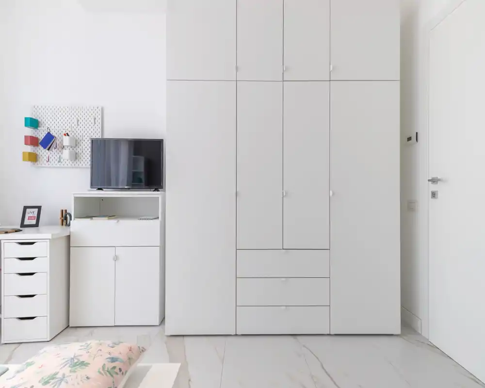 Coin chambre lumineux et minimaliste avec oreiller et couette clairs visibles, grand placard blanc intégré et petit espace bureau avec TV — moderne, propre et aéré.