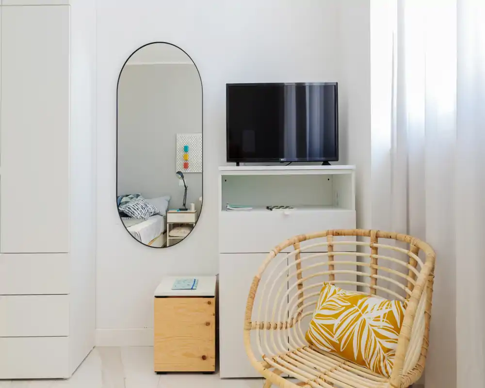 Bright, minimalist bedroom corner with a rattan chair, decorative cushion and a mirror reflecting the bed; white cabinetry and a TV create a clean, modern feel.