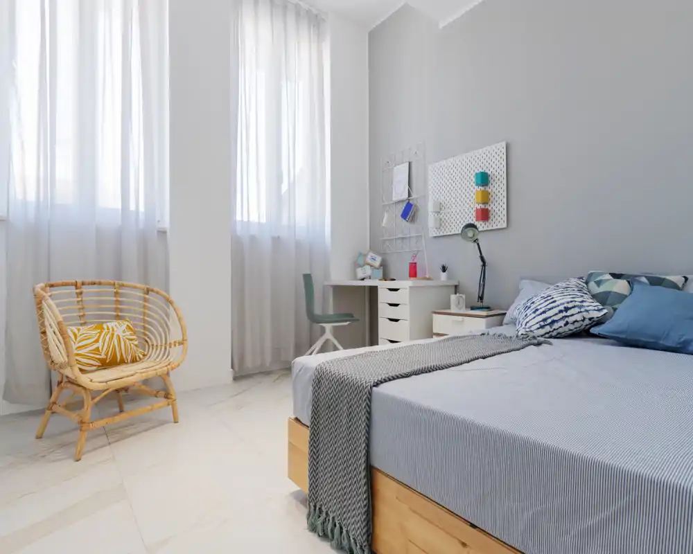 Bright, modern bedroom with a neatly made bed, soft textiles and a rattan accent chair by large sunlit windows — styled for comfort and relaxation.
