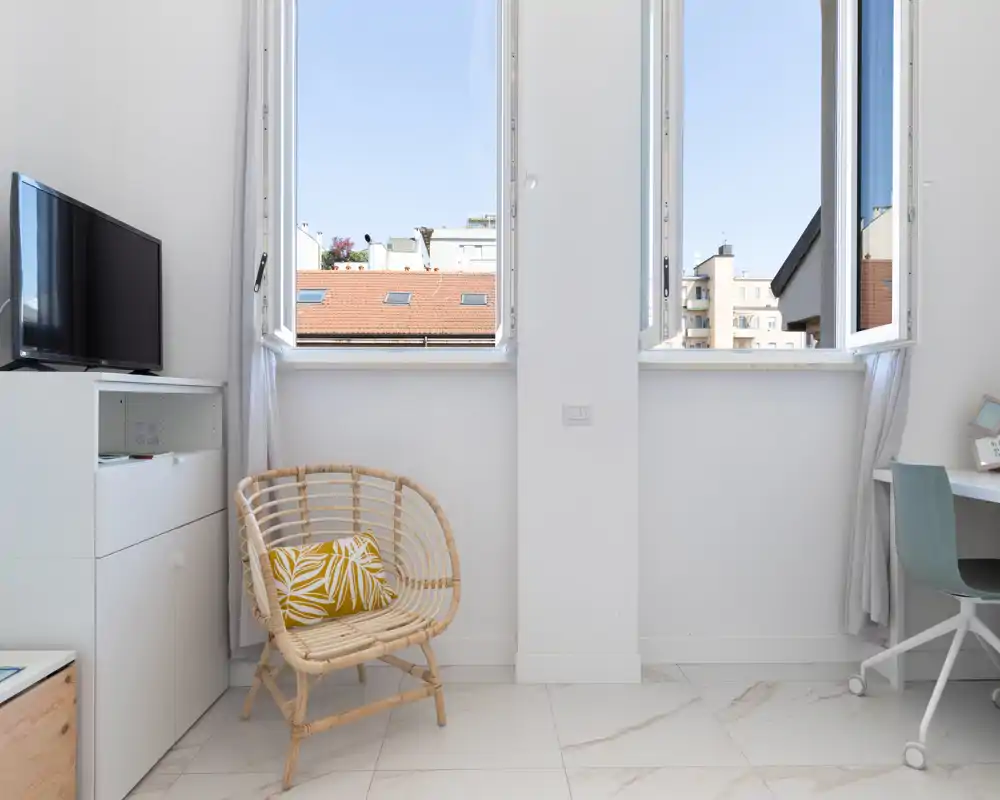 Bright, minimalist living area with two large open windows, a rattan chair with a patterned cushion, TV cabinet and a small desk — ideal for a compact, airy apartment.