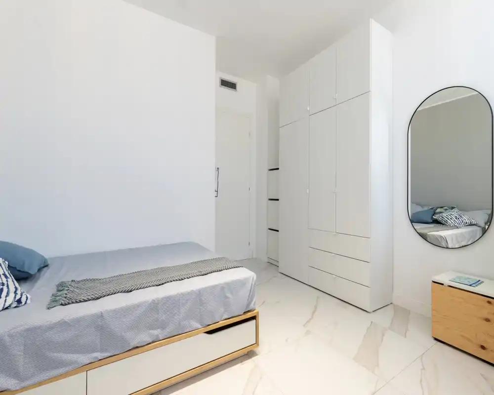 Bright, modern bedroom with a neatly made single/double bed, built-in wardrobe and a wall mirror — clean minimalist styling suitable for short-term rentals.
