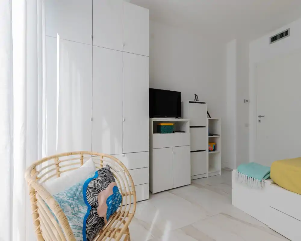 Bright, modern bedroom corner with built-in white wardrobes, a small TV unit and a bed partially visible on the right; cozy accents include a wicker chair, colourful cushions and a throw.