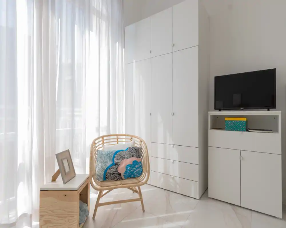 Bright, minimal living area with a rattan accent chair, soft cushions and white storage units; large sheer curtains let in abundant natural light.
