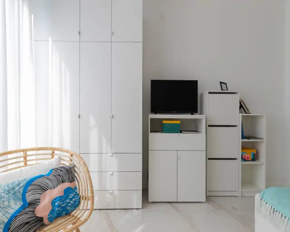 Bright, minimal living area with white storage units, a TV on a compact cabinet and a rattan chair with colorful cushions — airy and well lit.