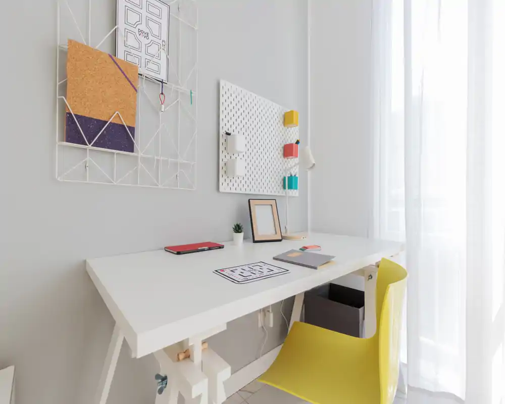 Bright, minimal home workspace with a white desk, yellow chair, pegboard organizer and soft natural light through sheer curtains.