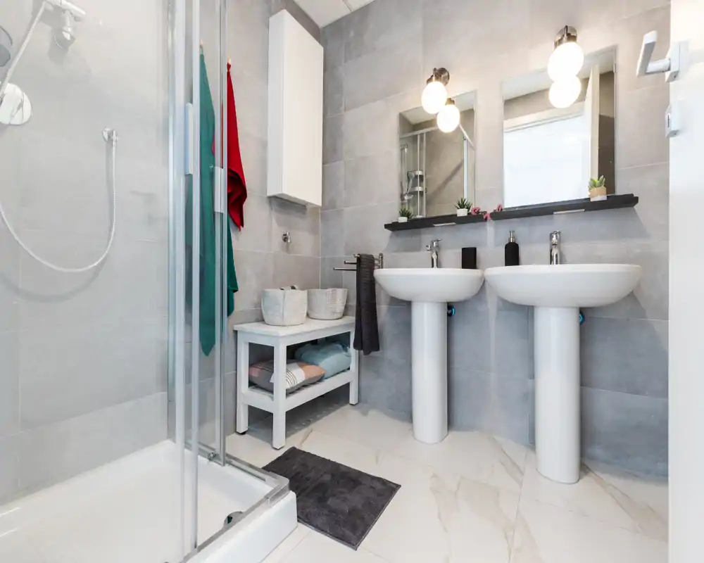 Bright, modern bathroom with a glass shower enclosure, twin pedestal sinks and contemporary fixtures. Neutral gray tiles and white fittings create a clean, minimalist look.