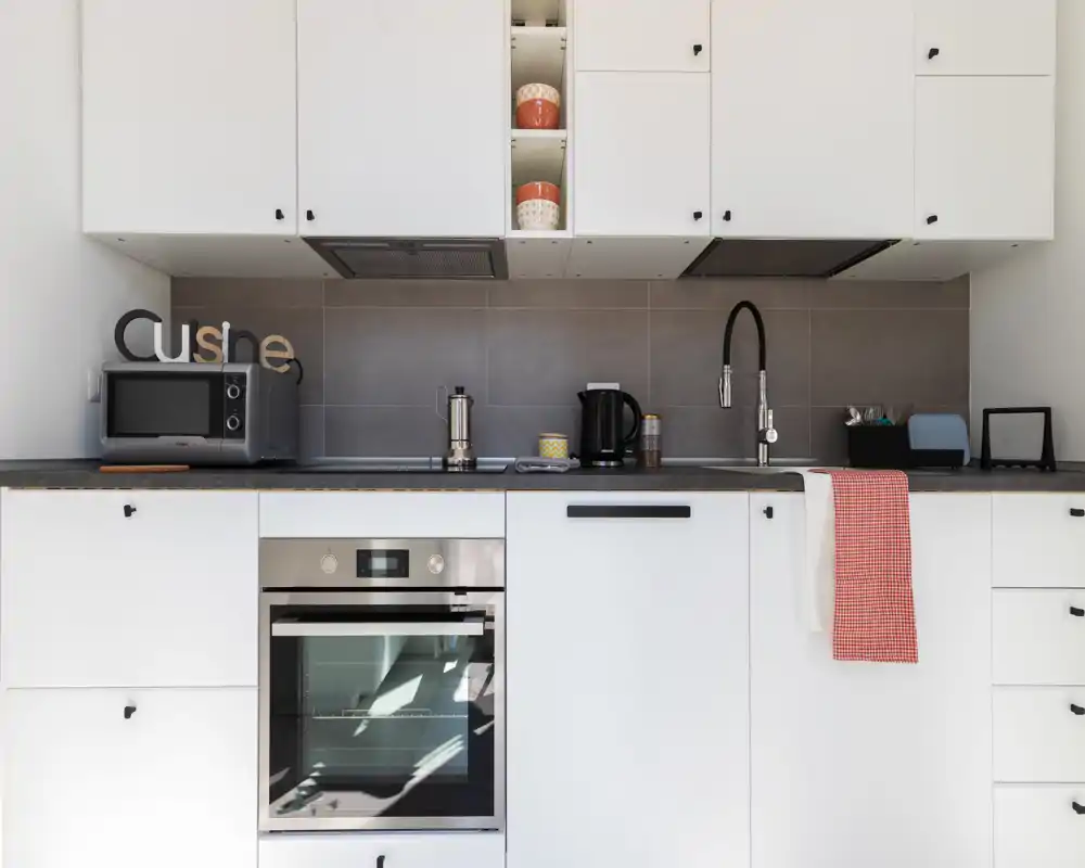 Modern compact kitchen with white cabinetry, built-in oven, microwave and a sleek black faucet; tidy countertop with kettle and small decorative items.