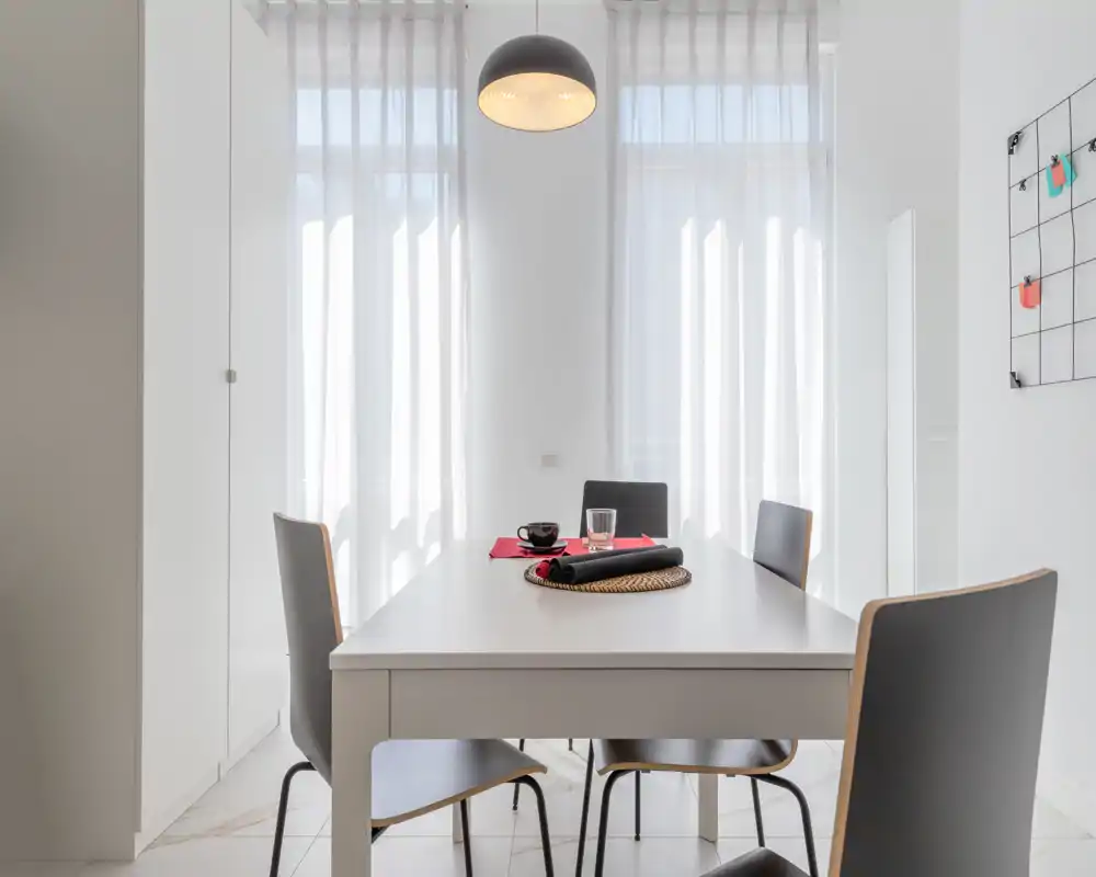 Minimal, bright dining room with a white table, four modern chairs and a hanging pendant lamp; natural light filters through sheer curtains for an airy feel.