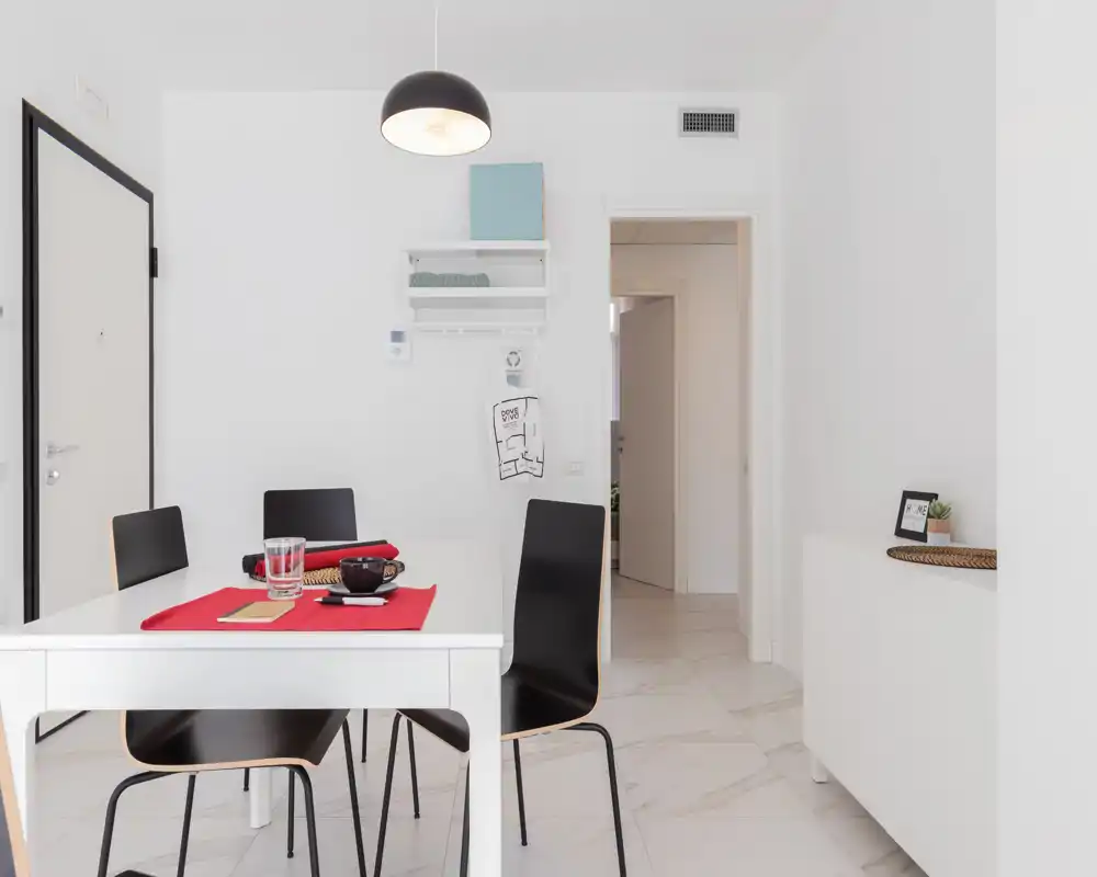 Bright, minimalist dining area with a white table, four black chairs and a simple red table runner; clean lines and marble-look floor make this space feel modern and airy.