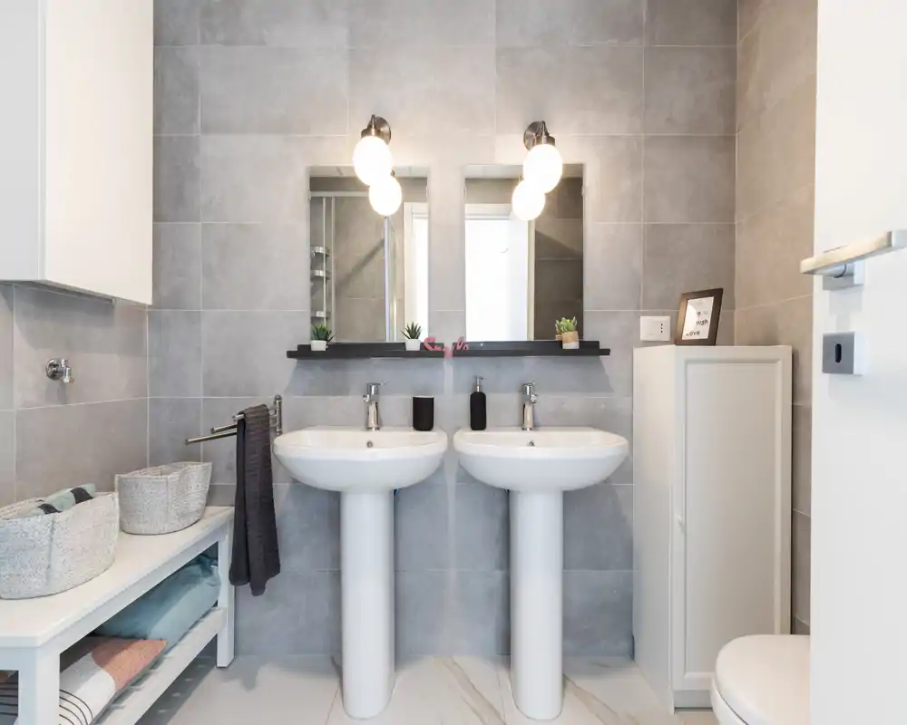 Bright modern bathroom with twin pedestal sinks, wall-mounted mirrors and contemporary gray tiles; organized shelving and soft textiles create a clean, functional look.