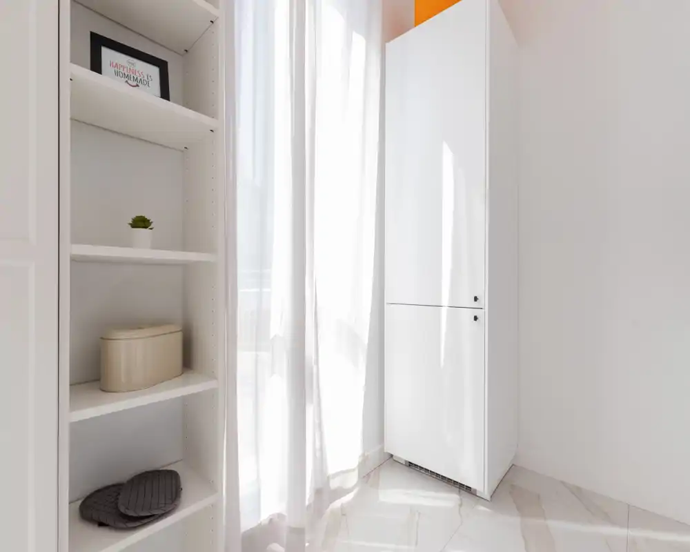 A bright, minimal storage corner with built-in shelving and a tall white cabinet next to sheer curtains letting in natural light. The space feels clean and functional for storage or as a small wardrobe area.