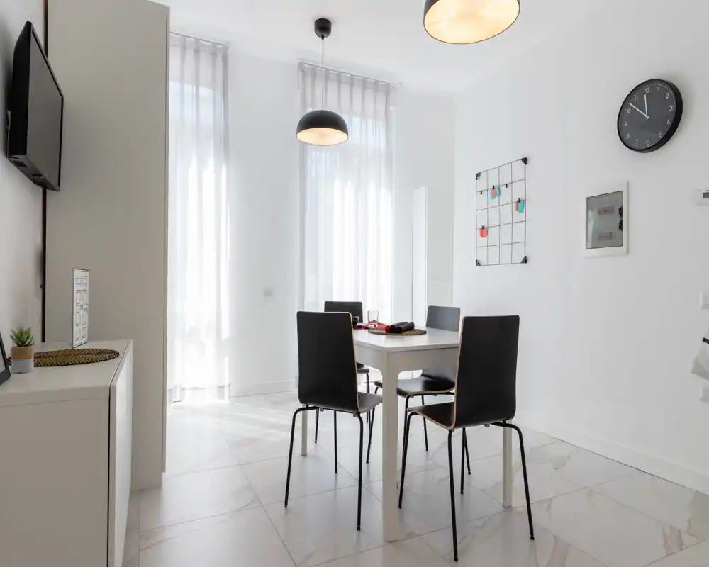 Bright, modern dining area with a white table and four black chairs set under pendant lights, tall sheer curtains and minimalist decor.