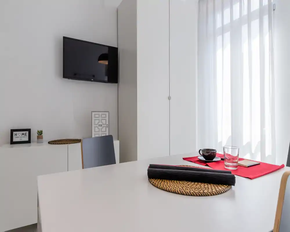 Minimal, bright living/dining corner with a white table, placemat and cup, wall-mounted TV and tall wardrobe; neat, modern and simply styled.