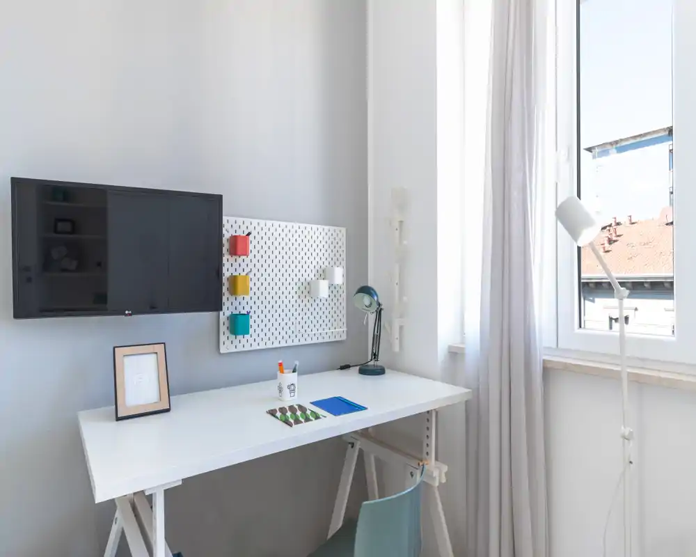 Angolo lavoro moderno e luminoso con scrivania bianca, lampada da tavolo, pannello portaoggetti e TV a parete — perfetto per studio o lavoro con molta luce naturale.