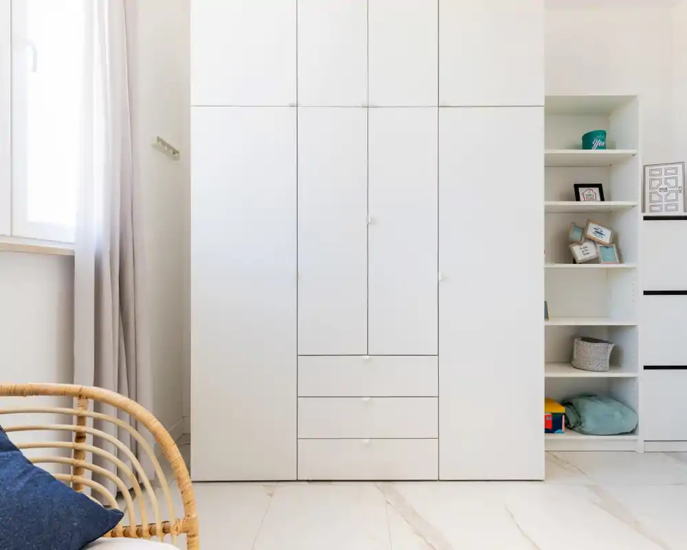 Angolo di camera luminoso e minimal con un grande armadio a muro bianco, scaffalature con oggetti decorativi e una poltrona in midollino con cuscini.