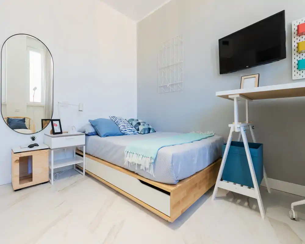 Camera da letto luminosa e moderna con letto matrimoniale ben rifatto, tessuti blu e una piccola scrivania con TV a parete — ideale per un soggiorno confortevole in città.