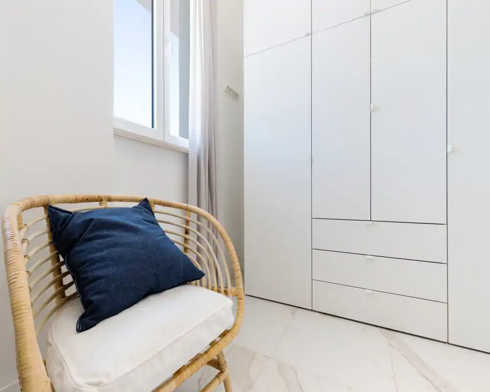 Angolo di camera luminoso con armadi bianchi a muro e una poltrona in rattan con cuscini, illuminato da luce naturale da una finestra.