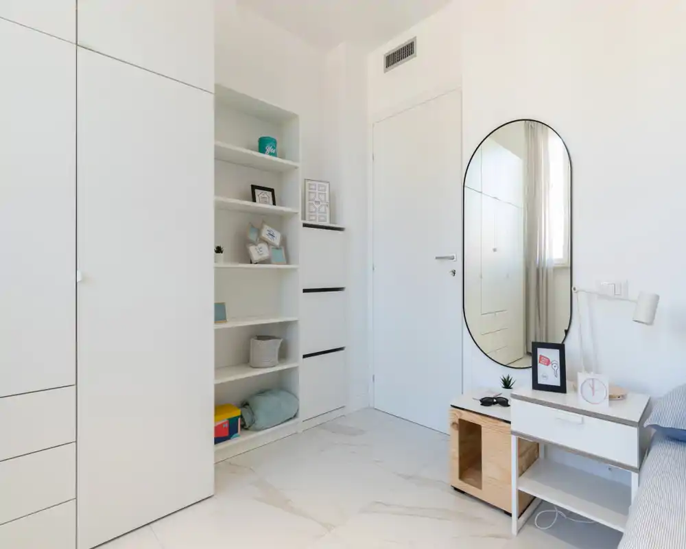 Angolo camera luminoso e minimal con armadi a incasso, scaffali e un piccolo comodino; una porzione del letto è visibile sul lato destro. Finiture bianche e luce naturale donano un aspetto moderno e fresco.