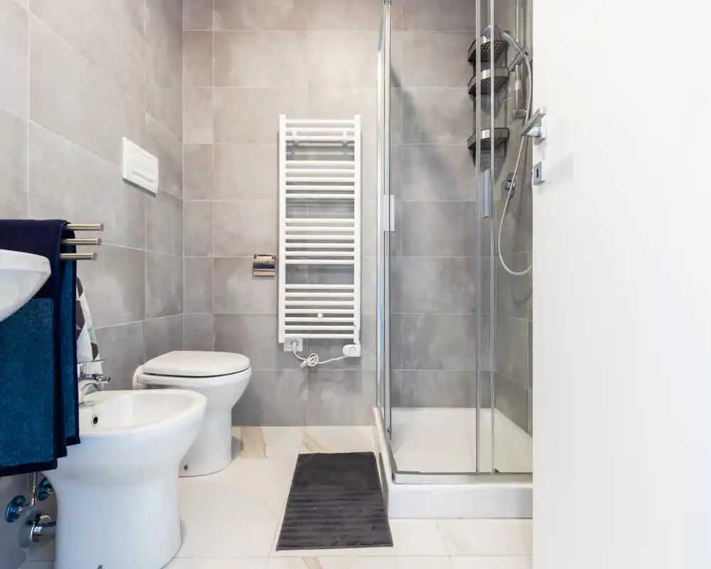 Bagno moderno e compatto con box doccia in vetro, wc, bidet e termoarredo a parete; piastrelle grigie neutre e sanitari bianchi per un aspetto pulito e contemporaneo.
