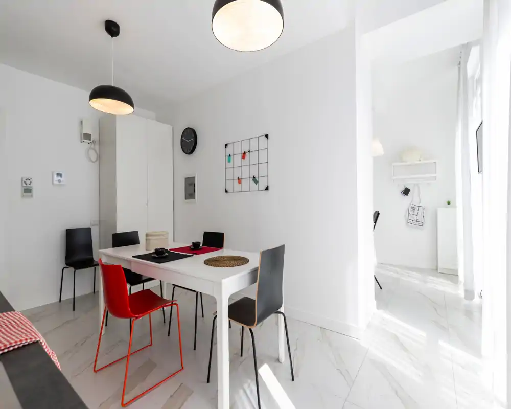 Zona pranzo luminosa e moderna con tavolo bianco, sedie di colori diversi e arredi minimal. Ampie finestre che assicurano molta luce naturale per un ambiente arioso e contemporaneo.