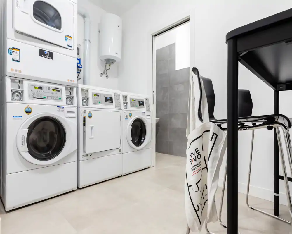 Locale lavanderia/utility luminoso e pulito con lavatrici e asciugatrici impilate e un piano di appoggio con sedie — ideale per convivenze o servizi condominiali.