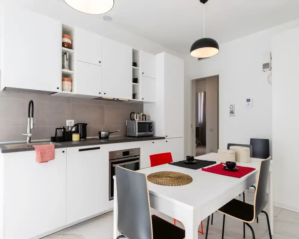 Cucina moderna bianca con elettrodomestici integrati, tavolo da pranzo apparecchiato e lampade a sospensione eleganti; ambiente luminoso, pulito e funzionale ideale per affitti brevi.