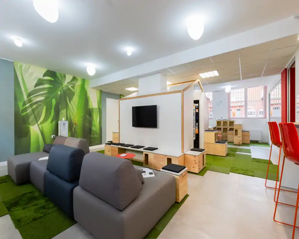 Area comune moderna e luminosa con divani modulari, TV a parete, un murale verde e posti a sedere informali — ideale per rilassarsi o socializzare.