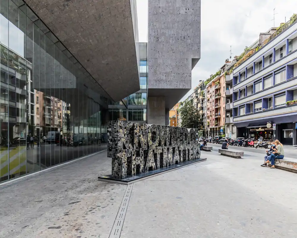 Piazza esterna pubblica davanti a un edificio moderno, con una scultura a lettere e panchine sul marciapiede.