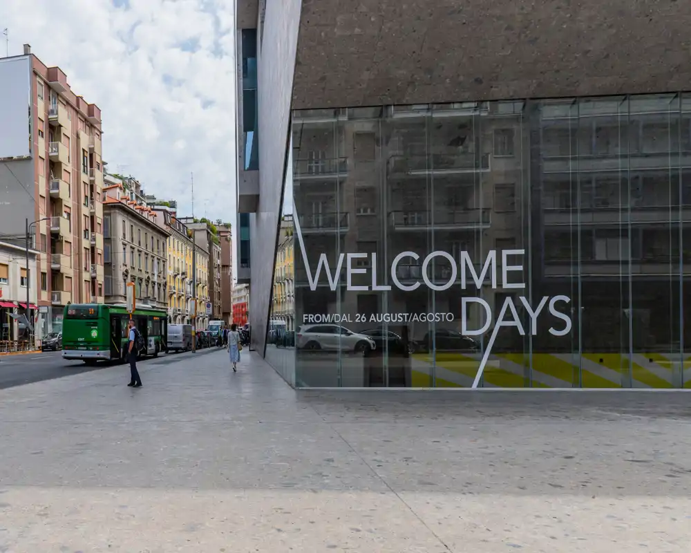 Esterno urbano con facciata moderna e grandi vetrate, strada visibile con autobus e pedoni; luce diurna brillante e superfici riflettenti creano una scena di piazza chiara e accogliente.