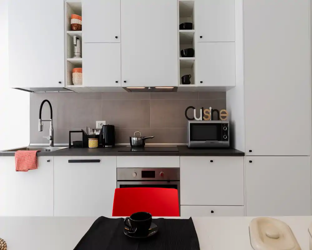 Cucina moderna e luminosa con mobili bianchi, forno integrato, microonde e lavello con rubinetto nero; dettagli curati come la sedia rossa, lo strofinaccio e la tazza in primo piano.