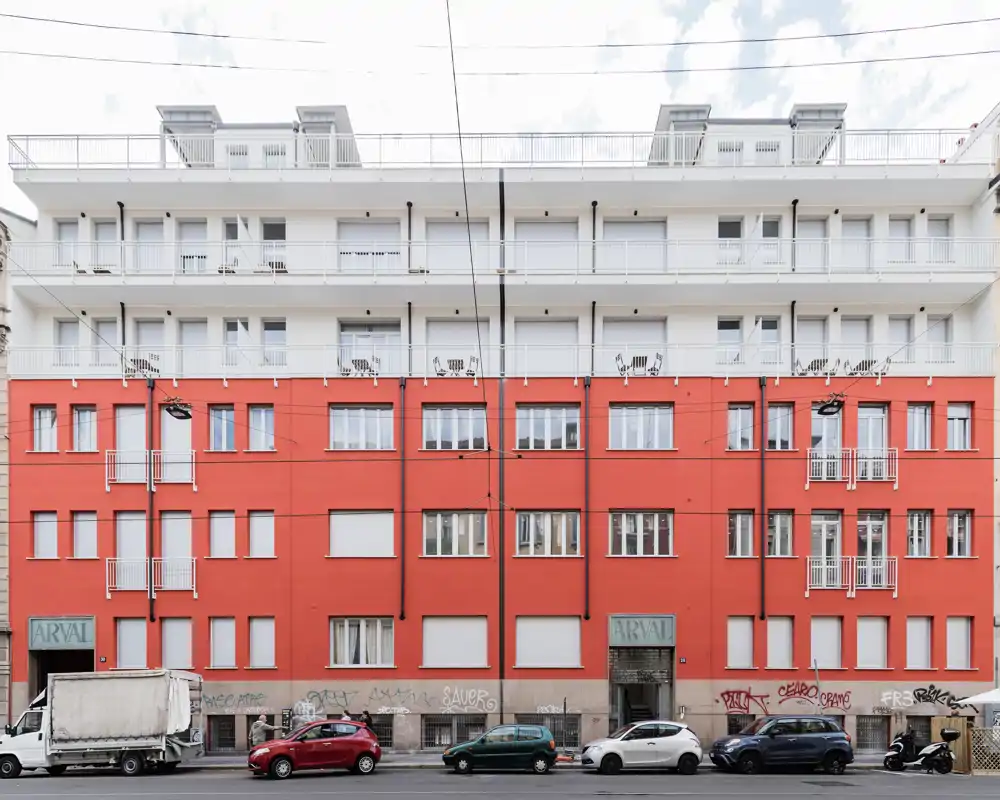 Vista esterna di un edificio residenziale a più piani con facciata rossa, balconi e strada con auto parcheggiate davanti. Buoni dettagli e luce naturale, adatto per presentare l’edificio o unità con affaccio sulla strada.