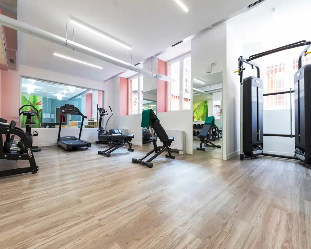 Sala fitness luminosa con tapis roulant, panca regolabile e macchine a cavo su pavimento effetto legno — piccolo spazio palestra ben attrezzato per gli allenamenti.