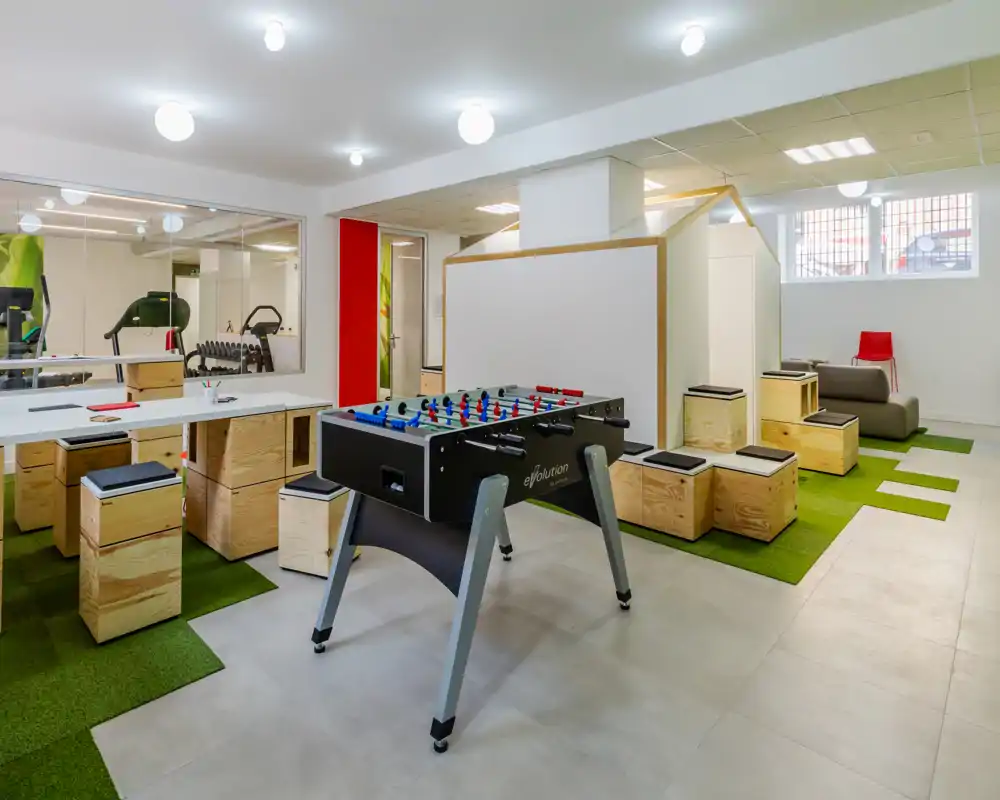 Spaziosa area ricreativa comune, luminosa e moderna, con calcio balilla, sedute modulari in legno e tavoli da lavoro: perfetta per socializzare e lavorare in modo informale.